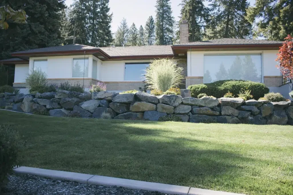 Greenscape project photo — a tiered basalt boulder retaining wall holding a planted bed above a clipped lawn, in front of a mid-century home
