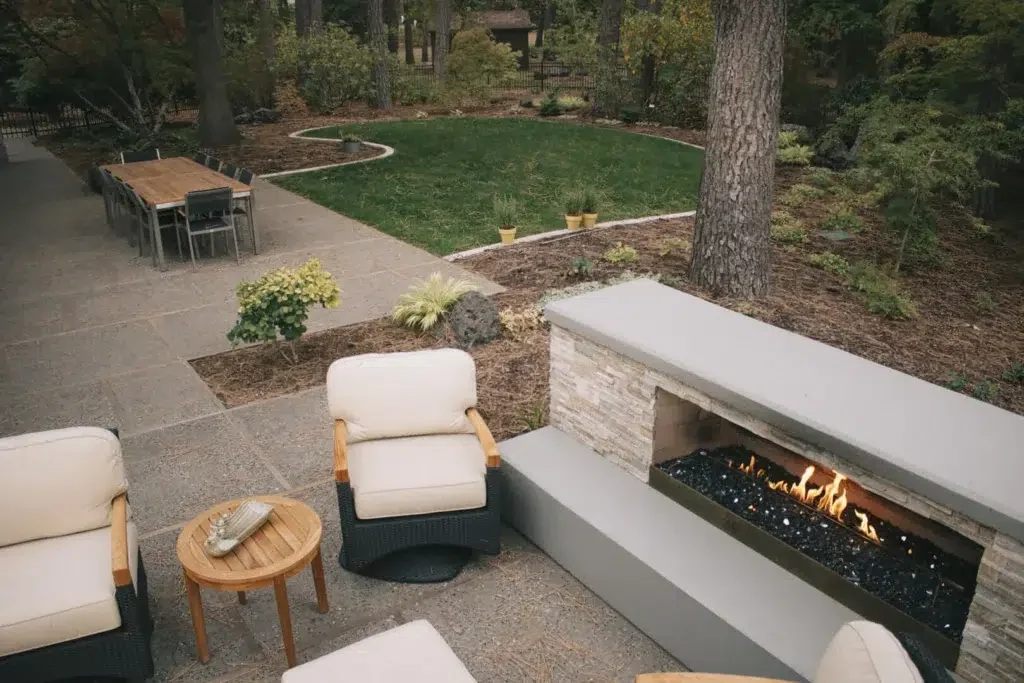 Greenscape project photo — a flagstone patio with a long linear gas fireplace, cushioned seating and a dining table under mature pines