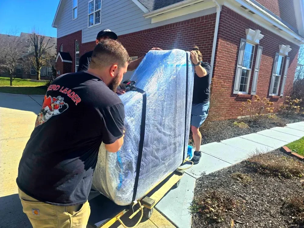 Immediate Movers project photo — two movers easing a shrink-wrapped upright piano on a piano board down a driveway