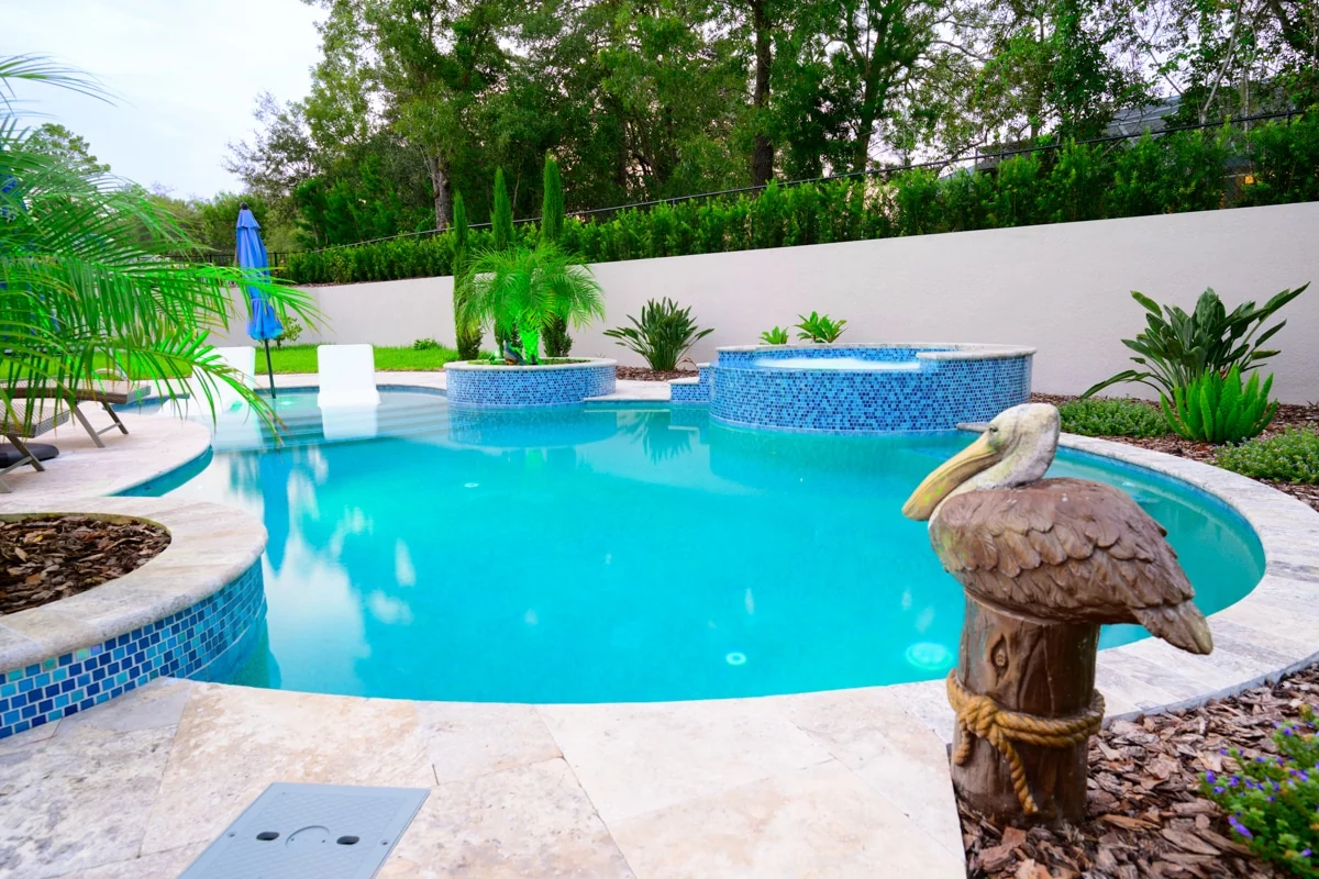 Nic’s Pool Construction project photo — a freeform pool with two raised blue mosaic spas and travertine coping, a carved pelican on a piling in the foreground