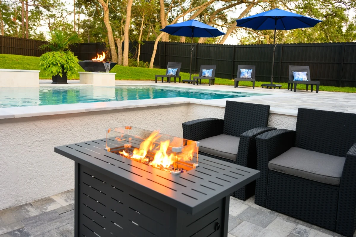 Nic’s Pool Construction project photo — a lit fire table and wicker seating in the foreground with the pool, fire bowl and blue umbrellas behind