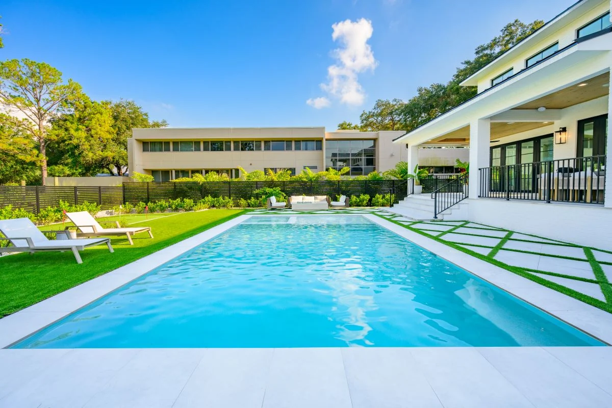 Nic’s Pool Construction project photo — a rectangular pool with clean lines, light stone pavers and turf inlays beside a white modern house