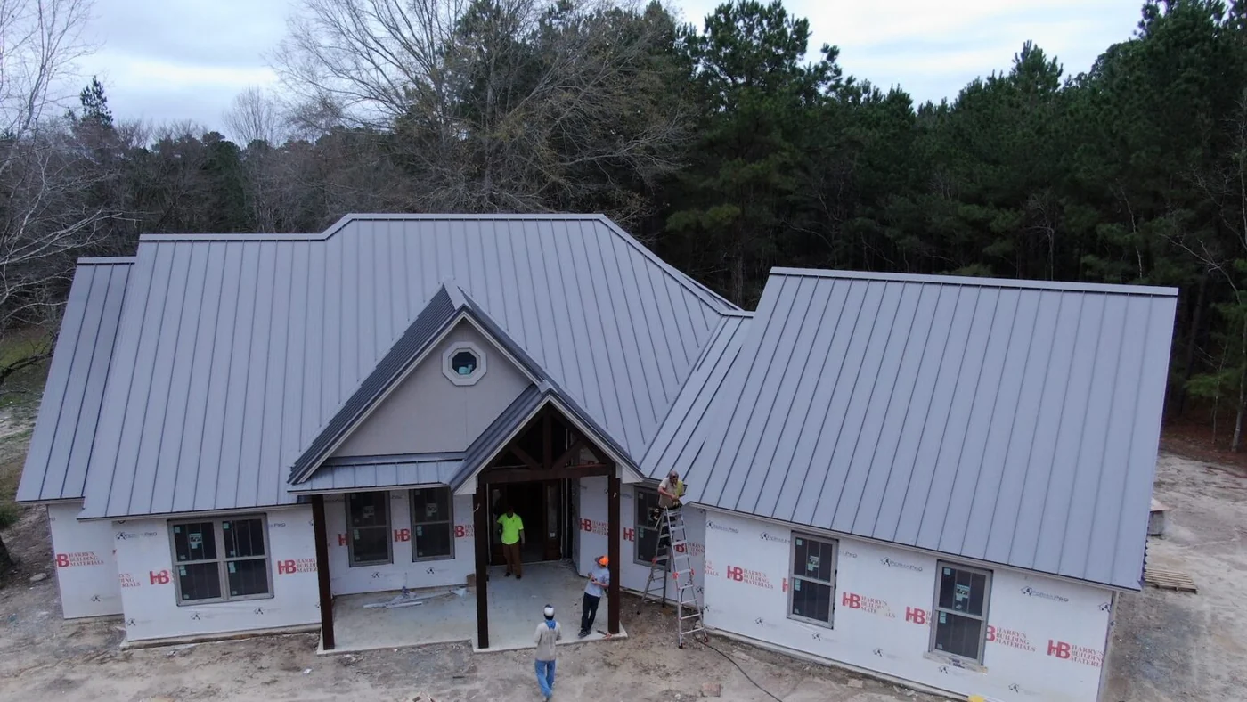 Roof Ryders project photo — a drone view of a new build still in housewrap, with the crew on ladders finishing a grey standing-seam metal roof