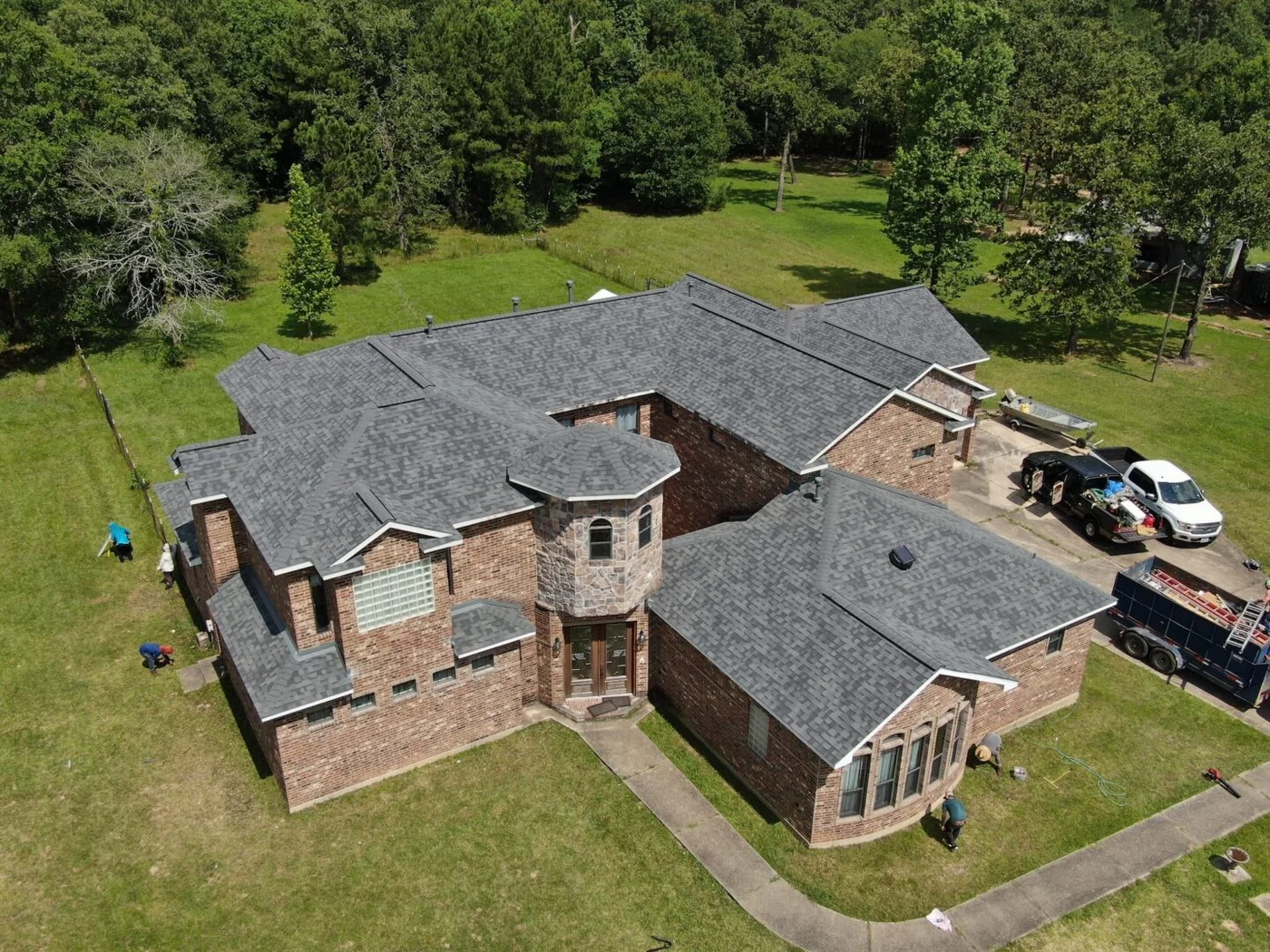 Roof Ryders project photo — a drone view of a large brick home in Lufkin, Texas with a new grey shingle roof, the crew and trailer still on site