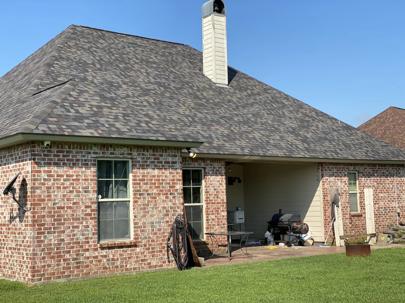 Roof Ryders project photo — a ground-level view of a red brick house with a new weathered-wood shingle roof and a white chimney against a clear sky